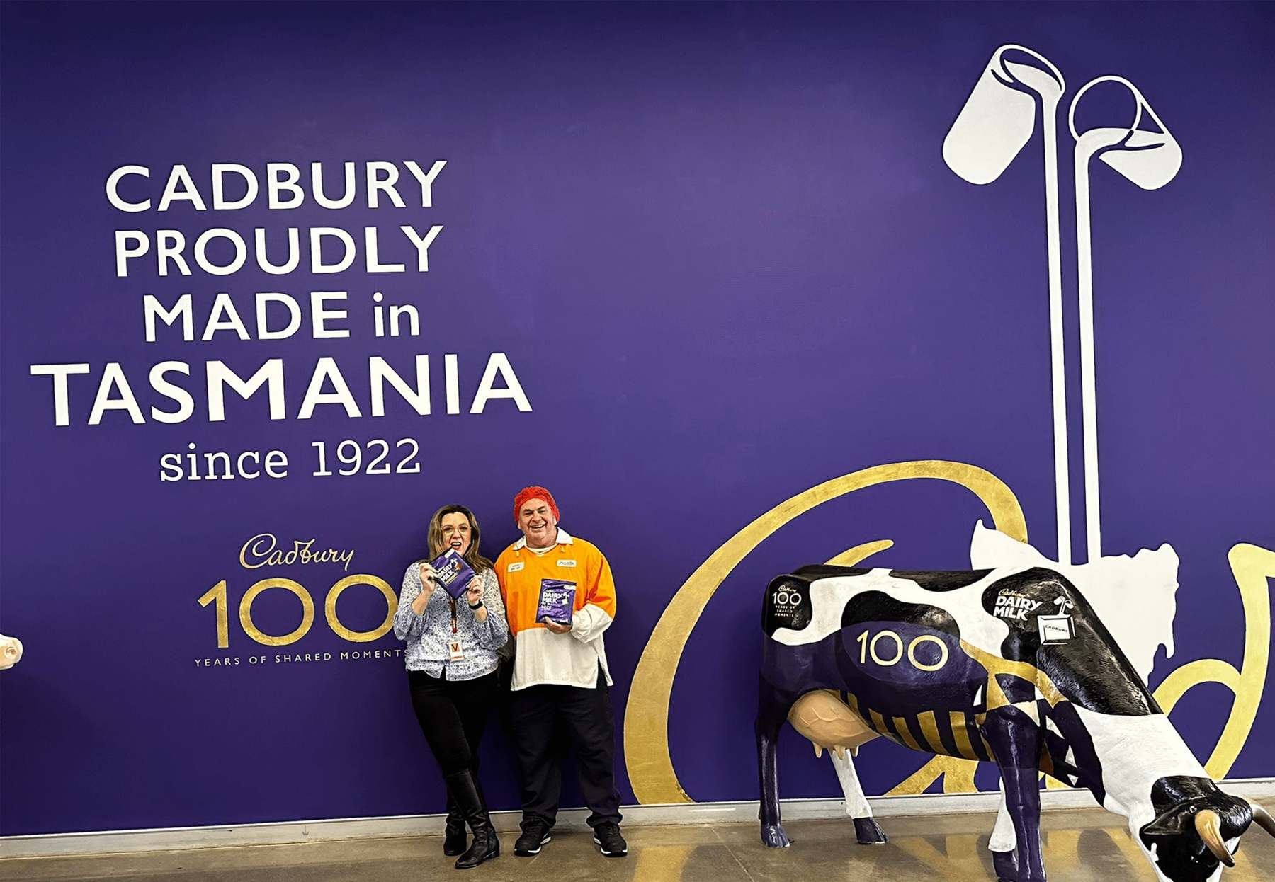 Tammy stands in front of a giant purple wall at Cadbury's with a man in a hairnet and an orange shirt. They are both holding chocolate.