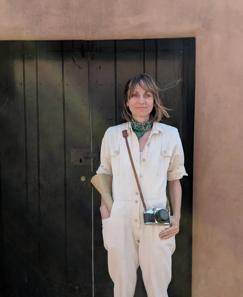 A woman in a white jumpsuit with a green bandanna around her neck stands in front of a dark wooden door wearing a camera